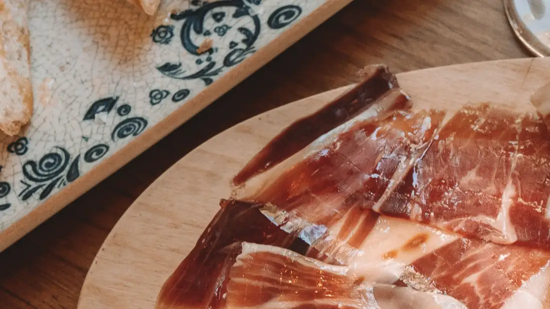 Jamón cortado a cuchillo sobre tabla de madera, con pan con tomate en un plato de azulejo